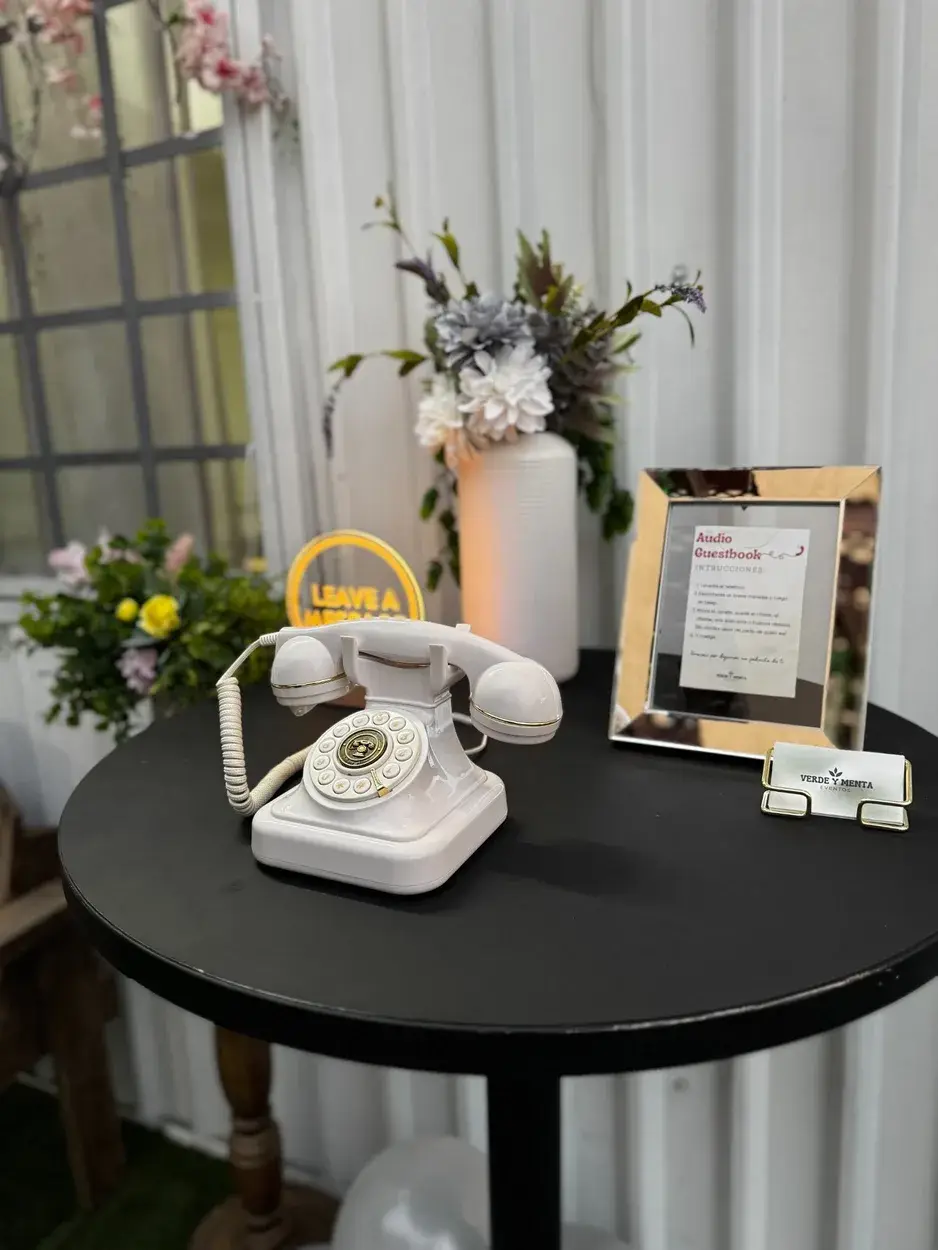 Teléfono retro blanco sobre mesa negra, con instrucciones en un marco dorado y flores al fondo